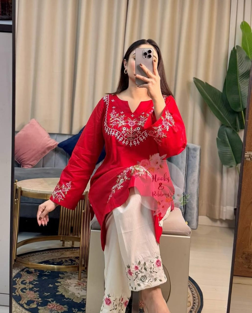 Woman taking a mirror selfie wearing a red embroidered top and white pants in a room.
